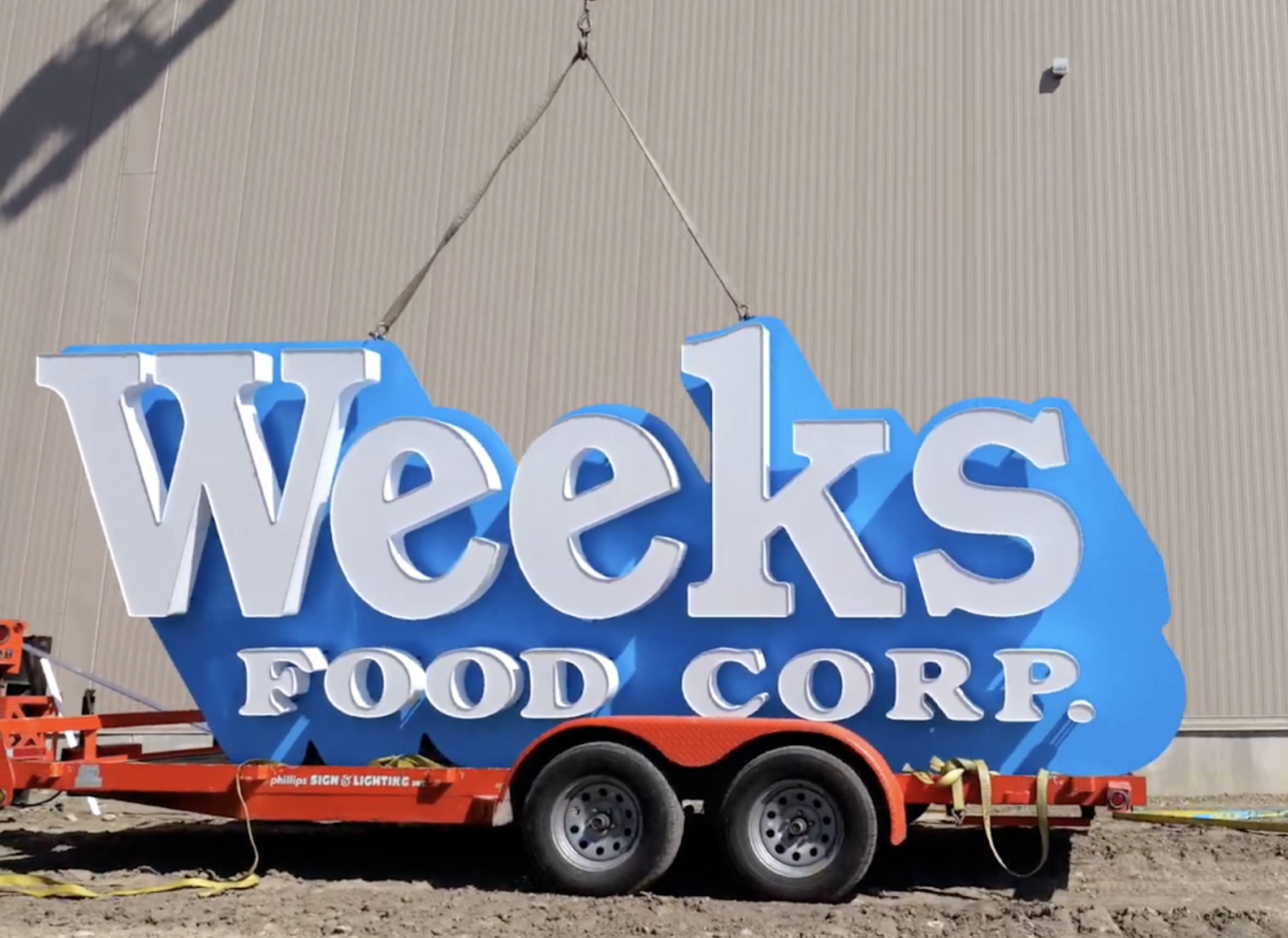 Large blue and white Weeks Food Corp. sign being hoisted by a crane off a flatbed trailer, ready for installation.